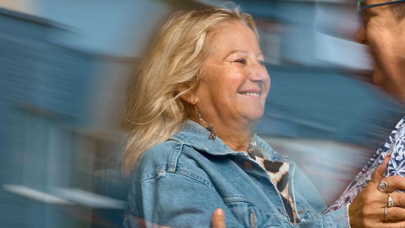 A smiling woman in a blue denim jacket reaches out to embrace another woman wearing glasses. Overlay text reads: "Expert patient and mentor."
