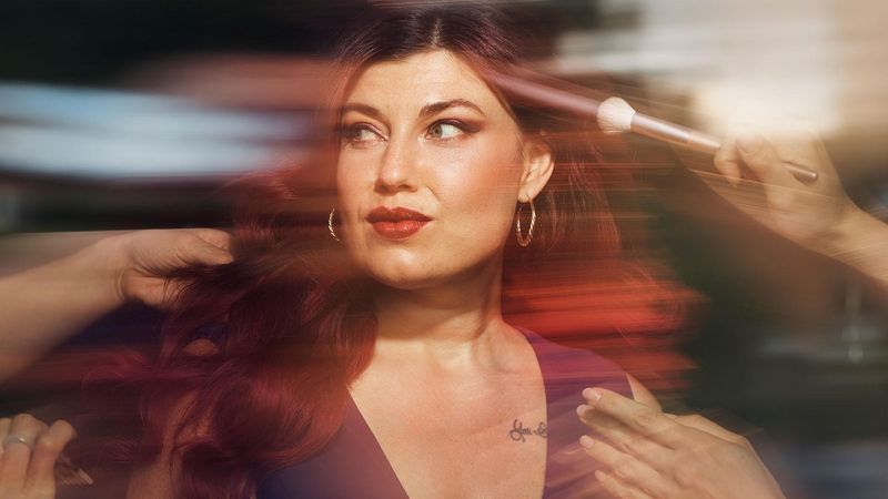 A glamourous woman wearing a purple dress is having her hair and makeup finished, she is looking to the side at somone as they fix her hair. Overlay text reads: "Model and role model."