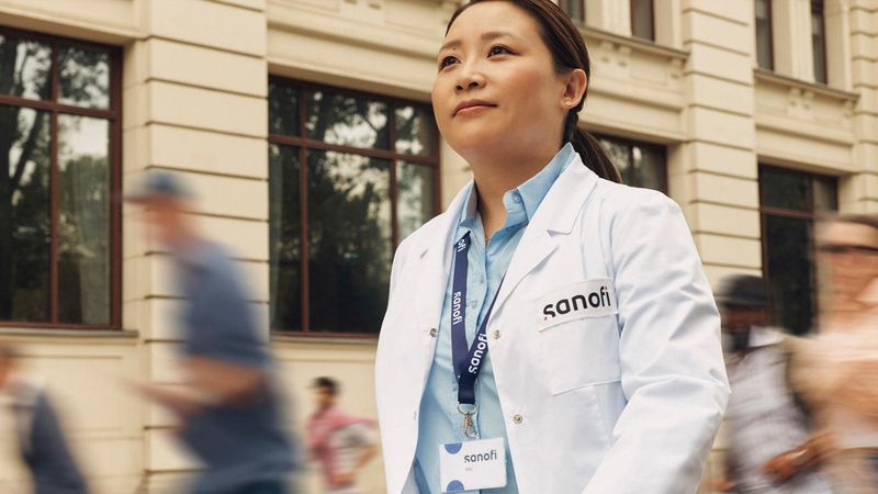 A woman in a white Sanofi lab coat and company badge outside of a building, moving forward while confidently and positively looking ahead. Several people are running alongside her in the background, they are blurred in motion.. Overlay text reads: "Working in Artificial Intelligence."